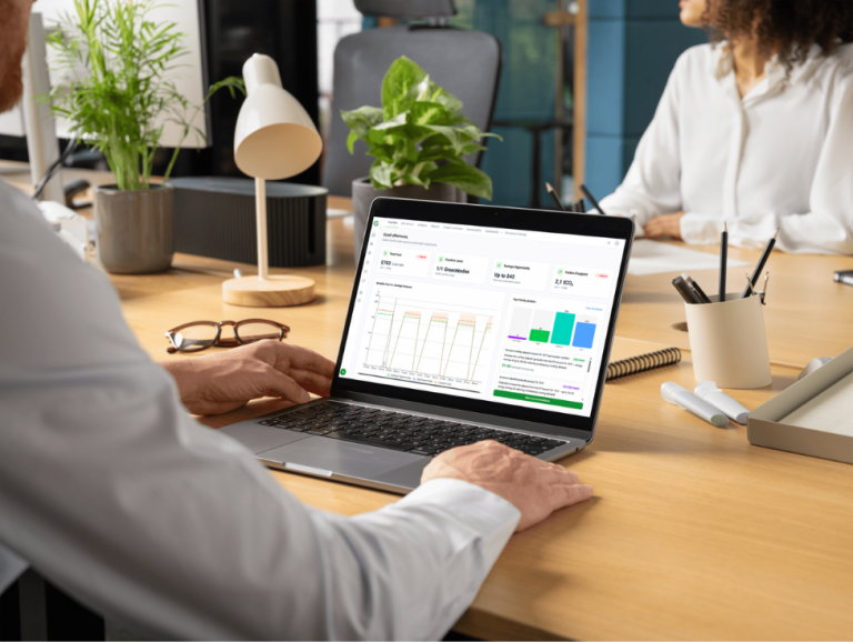 A man viewed over the shoulder, sitting at a wooden desk, focused on his laptop screen. The screen displays the GreenNode platform with a detailed dashboard showing KPIs such as 'Total Cost', 'Comfort Level', 'Savings Opportunity', and 'Carbon Footprint', along with charts for monthly forecasting and priority actions. He is working in a modern office with desk plants and a colleague in the background.