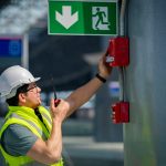 Building maintenance technician inspecting a wall-mounted system while holding a two-way radio, representing proactive facility operations and maintenance.