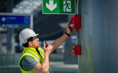 Building maintenance technician inspecting a wall-mounted system while holding a two-way radio, representing proactive facility operations and maintenance.